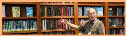 Image, a grey haired man points to a book among others on full wooden shelves.