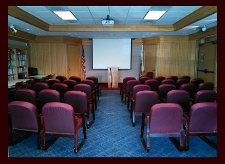 Photo showing a room full of padded chairs with a podium and  movie screen at the front.