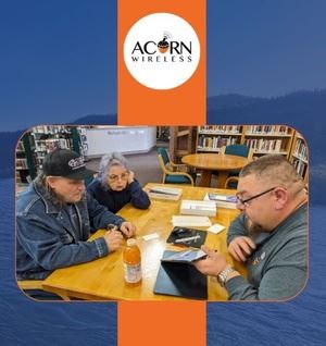 A large bearded man helps two people with tech questions at a library table.